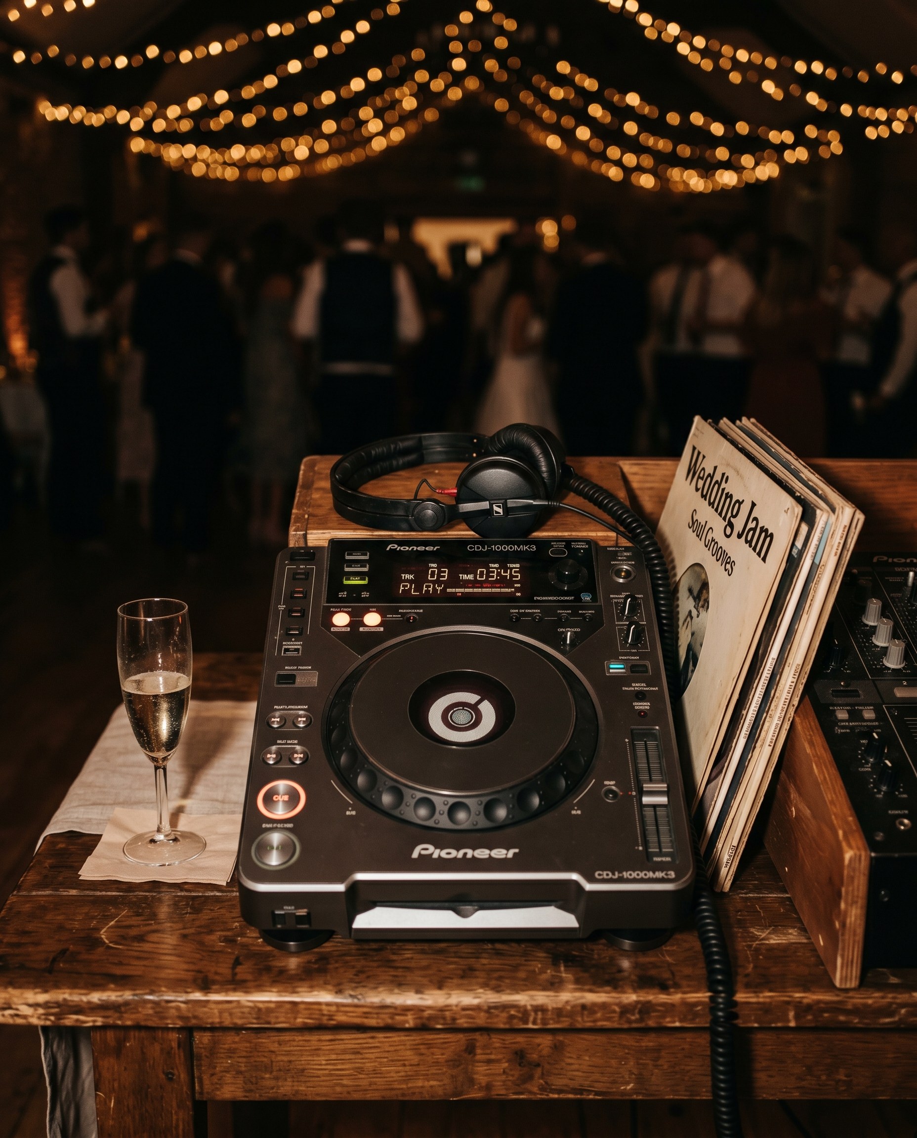 Detailed close-up photograph of vintage DJ equipment on a wooden table at a wedding reception, warm tungsten lighting, editorial style