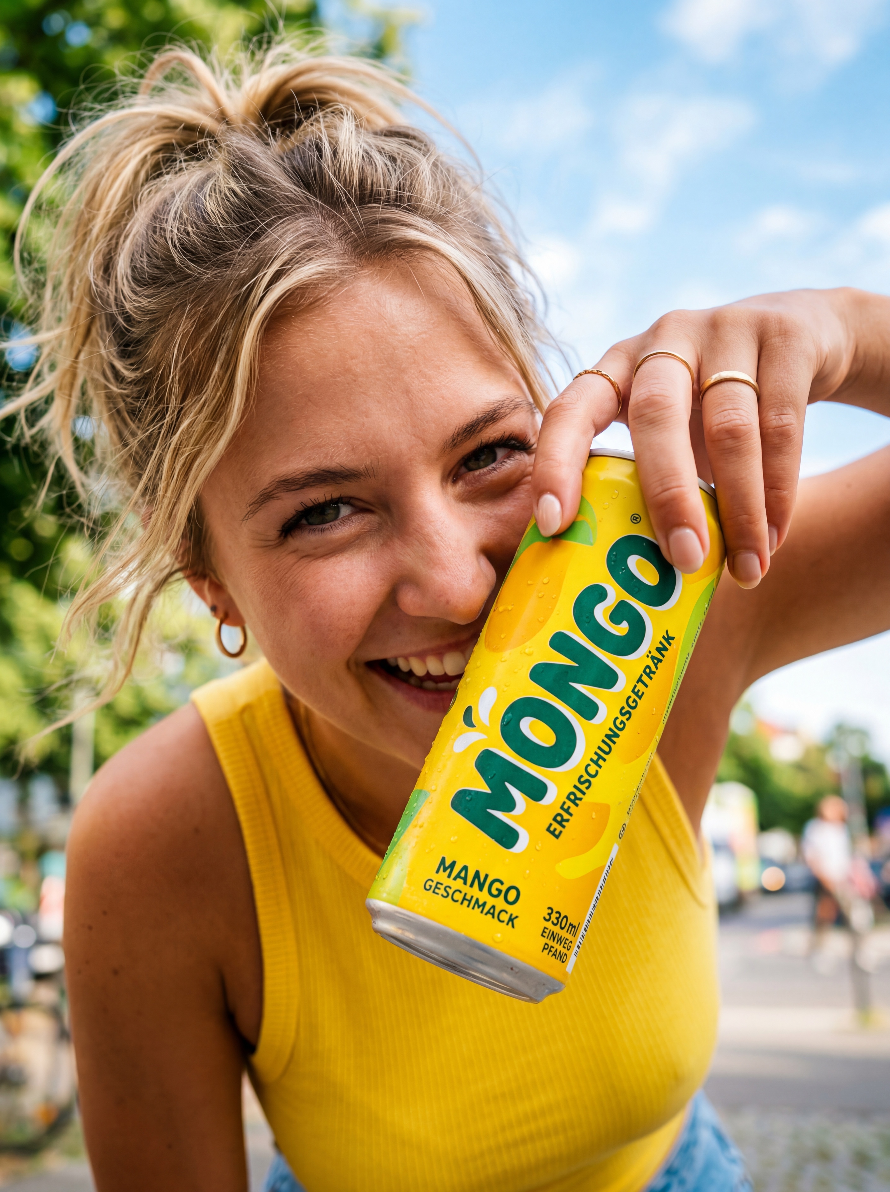 Nano Banana Pro sample image: Lifestyle product photography: young woman with messy blonde ponytail in sunny urban Berlin street, smiling while holding a yellow Mongo Mango energy drink can to her face, natural sunlight, candid commercial advertising style, ultra-realistic skin texture